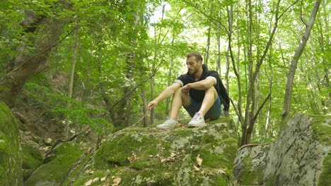 Thoughtful-young-man-in-the-forest-is-sitting-on-the-rock.
